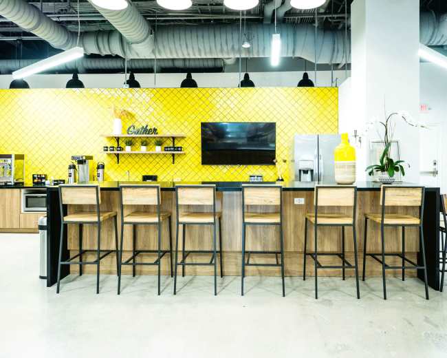 A modern kitchen area with a yellow tiled wall, a large television, and eight wooden bar stools aligned at a dark countertop.