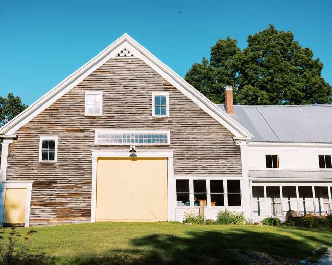 A weathered wooden barn with a yellow double door stands beside a white farmhouse under a clear blue sky.