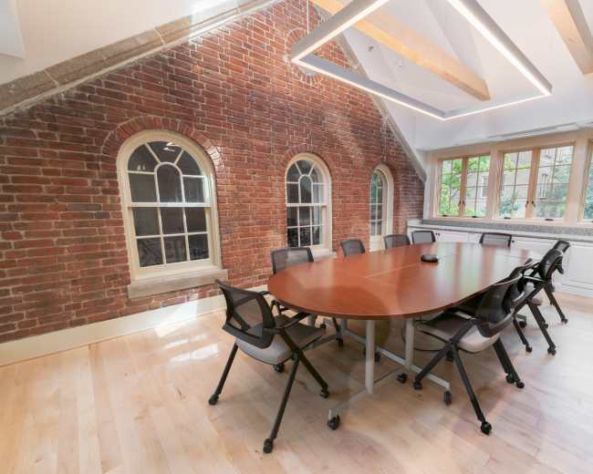 The image shows a modern conference room featuring a large wooden table surrounded by chairs, with exposed brick walls and large windows letting in natural light.