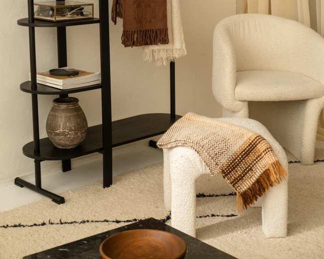A minimalist interior scene features a black shelving unit with various decorative items, a textured stool, and a cozy, light-colored chair.