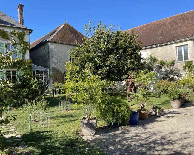 The image shows a garden with various plants and trees, surrounded by buildings with sloped roofs under a clear blue sky.