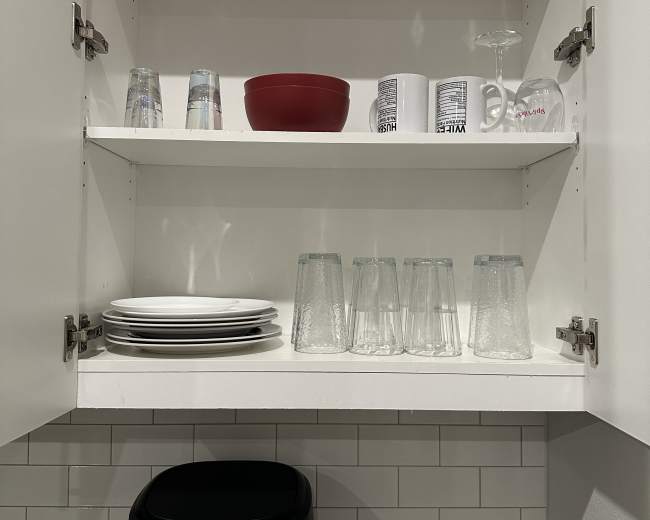 The image shows a kitchen cabinet with two shelves containing glasses, a red bowl, mugs, plates, and a black appliance on the counter below.