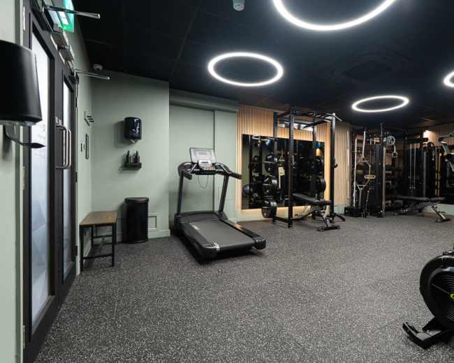 The image shows the interior of a modern gym featuring exercise equipment, including a treadmill and weightlifting stations, with circular overhead lights illuminating the space.