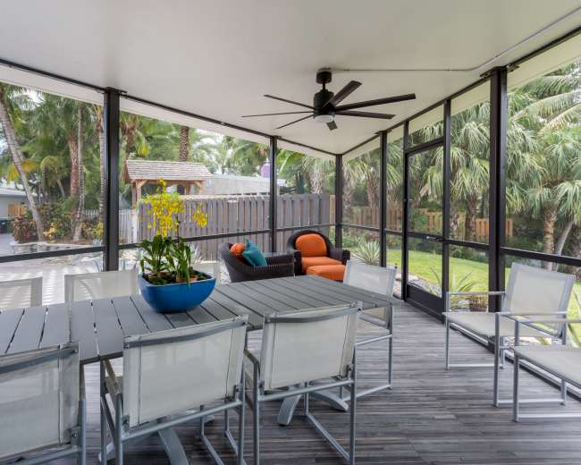 The image shows a screened-in patio with a dining table, chairs, and a seating area surrounded by palm trees and a landscaped yard.
