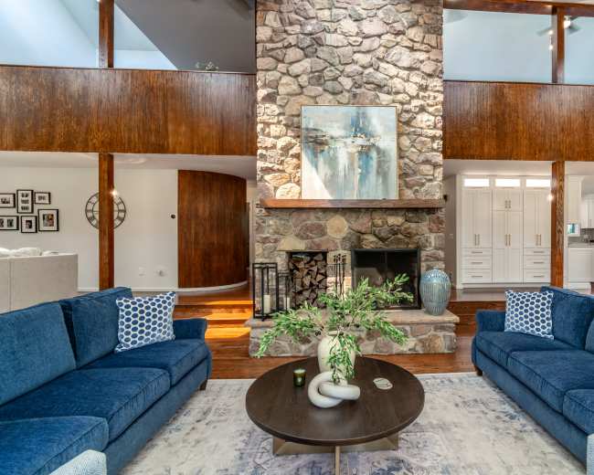 A living room features two blue sofas facing a stone fireplace with a large piece of artwork above it, surrounded by wood paneling and an open layout leading to a kitchen.