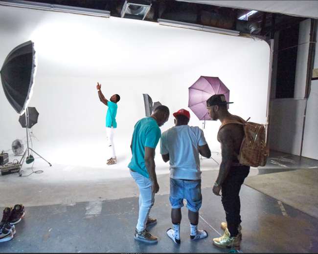 A group of three men is gathered in a photography studio with various lighting equipment and a plain white background.