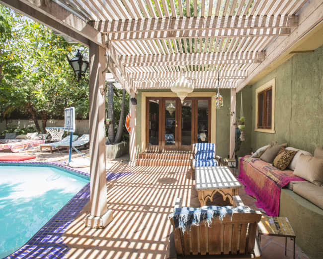 The image shows a patio area with a wooden structure overhead, a pool adjacent to it, and seating arranged along the side.