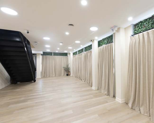 The image shows an empty indoor space with wooden flooring, beige curtains along the walls, and a staircase on the left side.