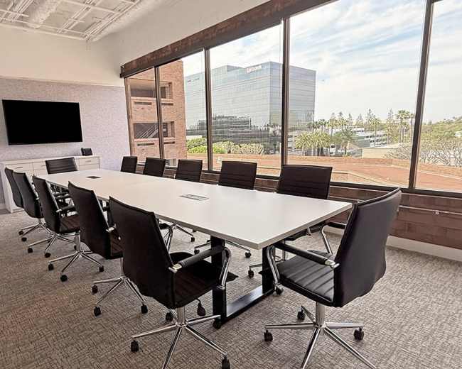 A modern conference room features a large white table surrounded by black swivel chairs, with large windows offering a view of an office building and the outdoors.