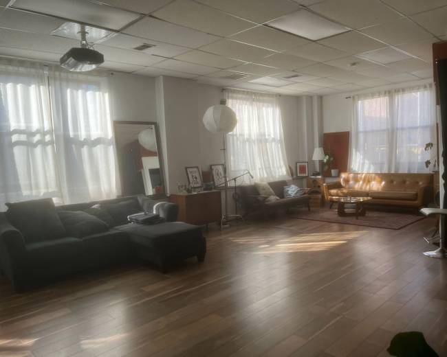 The image shows a spacious, well-lit living area featuring a large sectional sofa, a mid-century coffee table, and large windows with sheer curtains.