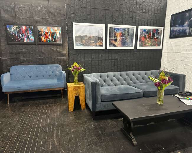 The scene features a stylish seating area with a blue tufted sofa, a matching loveseat, a wooden side table with a vase of flowers, and several framed photographs hanging on the wall.