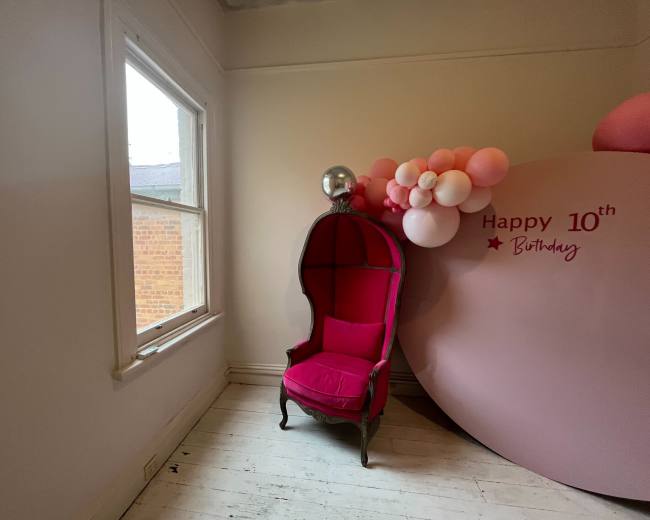 The image shows a pink chair in a room with a large round decoration that reads "Happy 10th Birthday" and a cluster of balloons nearby.