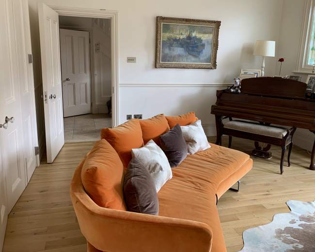A bright living room features an orange curved sofa with decorative pillows, a wooden piano, and a doorway leading to another room.