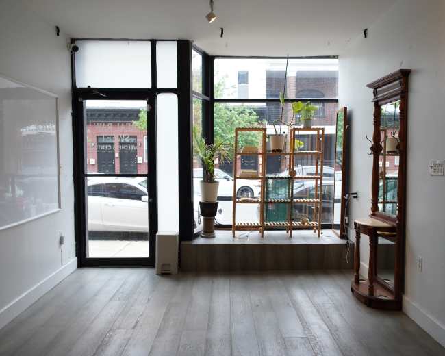 The image shows an empty commercial space with large windows facing a street, featuring a white wall, wooden flooring, and minimal furnishings including a display rack and a small table.