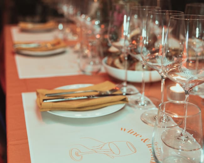 A long dining table is set with plates, wine glasses, and utensils, accompanied by decorative items and a tablecloth featuring wine-themed illustrations.