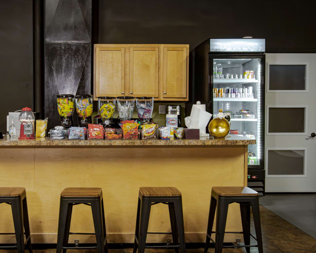 A modern snack bar features a countertop with various snacks and drinks, backed by a refrigerator and lockers.
