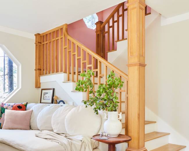 A sectional sofa is positioned next to a wooden staircase with a decorative railing and potted plant in a brightly lit room.