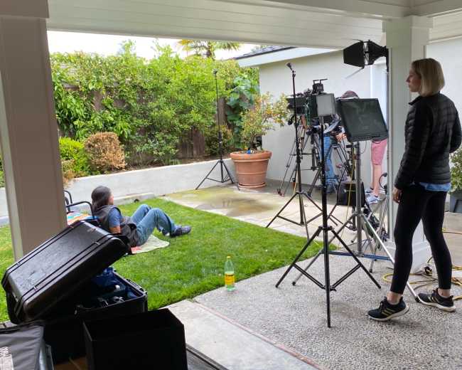A film crew sets up equipment in a backyard while a person sits on the grass.