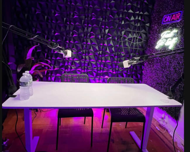 A podcast recording setup features a white table with three chairs, two microphones mounted on adjustable arms, and a textured wall illuminated with purple lighting.