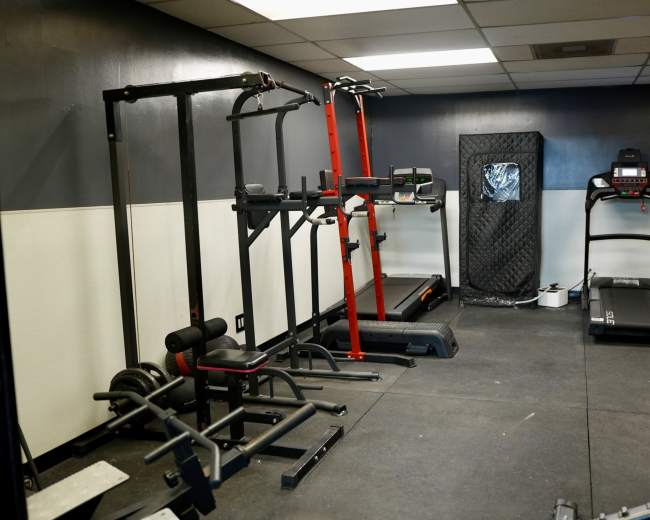 The image shows a small gym area equipped with various fitness machines and equipment, including a treadmill, pull-up bar, and weight rack.