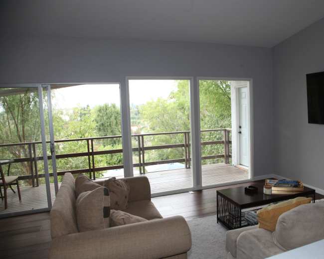 The image shows a living room with two sofas facing a coffee table and large windows that open to a balcony overlooking greenery.