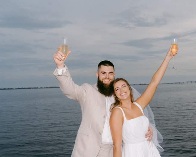 A well-lit path lined with palm trees leads to a body of water under a night sky.