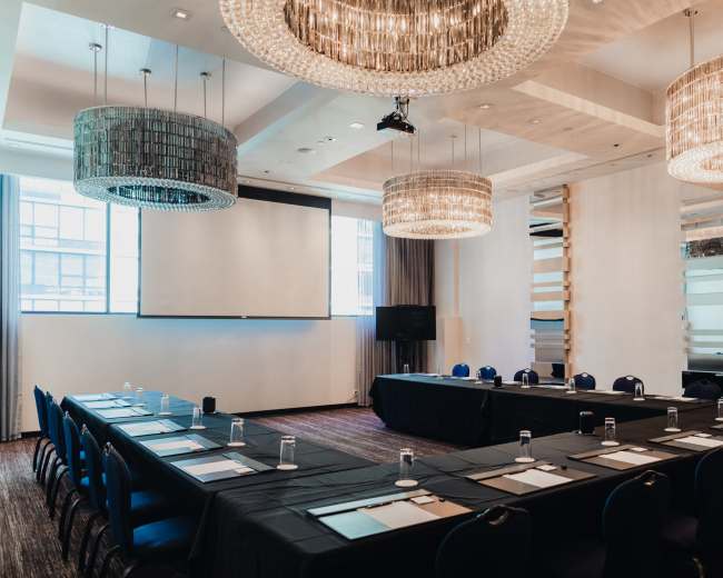 The image shows a modern conference room equipped with a long table arranged in a U-shape, illuminated by several elegant chandeliers.