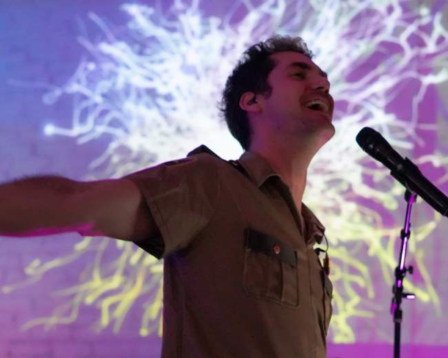 A performer with arms outstretched sings into a microphone against a colorful, abstract background.
