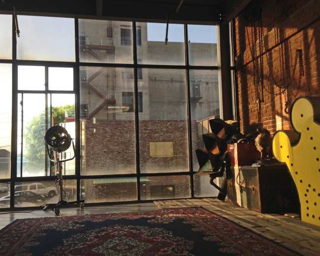 A spacious interior with large windows revealing a view of a building and parked cars outside, alongside a patterned rug and various objects on the floor.