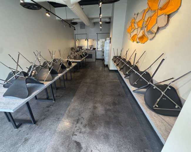 A series of tables with upside-down chairs are lined up in a modern, empty restaurant space.