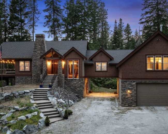 A large, rustic-style house with a stone facade and wooden accents is set amidst tall trees, featuring a three-car garage and a front porch with a staircase leading up to the entrance.