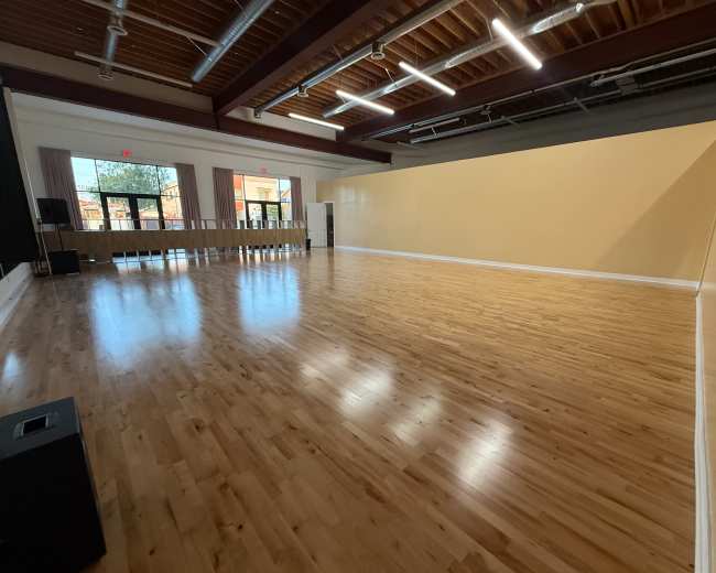 The image shows a spacious, empty studio with wooden flooring, large windows, and a plain yellow wall.