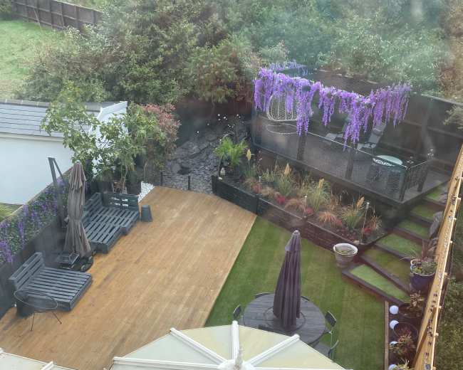 The image shows a landscaped backyard with wooden decking, garden furniture, and flowering plants under a purple wisteria trellis.
