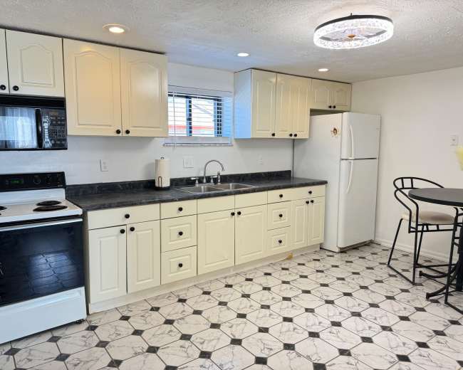 A kitchen with light-colored cabinets, a black countertop, a white refrigerator, an oven, and a small dining table with black chairs.
