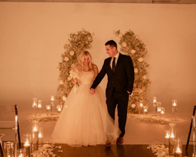 A bride and groom walk hand in hand down an aisle lined with candles and flower petals, flanked by floral arrangements.