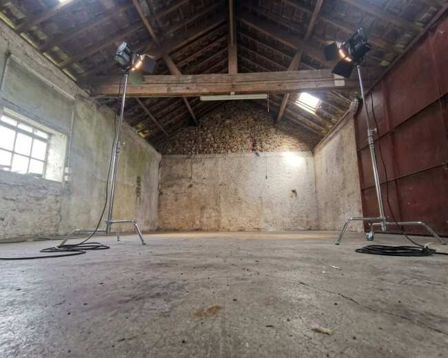 The image shows an empty, rustic interior of a room with bare stone walls and two studio lights positioned on stands.