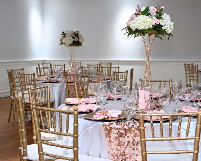 A banquet room is set with elegantly arranged tables featuring gold chiavari chairs, pink napkins, and floral centerpieces.