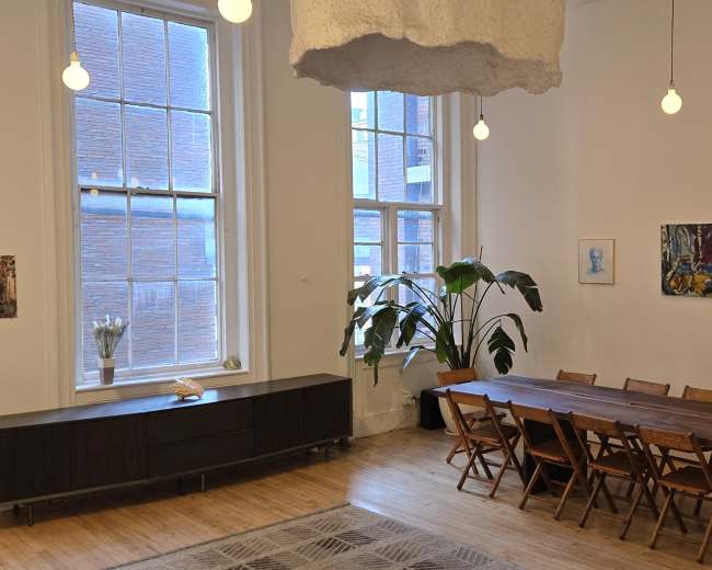 The image shows a spacious dining area with a long wooden table surrounded by folding chairs, two large windows allowing natural light in, and a few decorative plants and artworks on the walls.