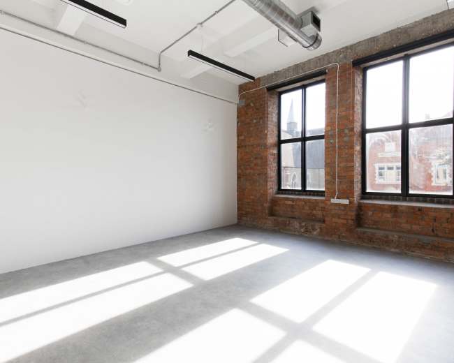 The image shows an empty interior space with a concrete floor and large windows allowing natural light to cast shadows on the walls.