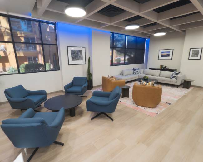 The image shows a modern lounge area with several chairs, a coffee table, and a sofa, illuminated by natural light from large windows.