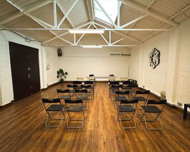 The image shows a spacious room with wooden flooring, featuring several rows of black folding chairs facing a small table and two additional chairs at the front.