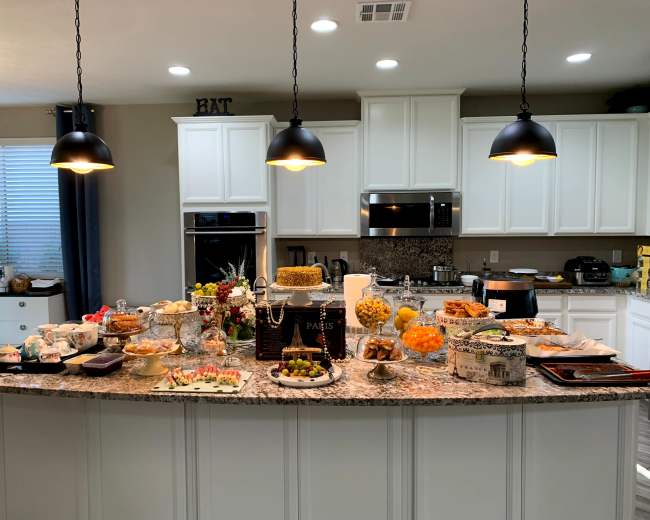 The image shows a kitchen island filled with a variety of dishes, desserts, and fruits, under hanging pendant lights.