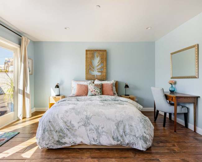 The bedroom features a queen-sized bed with a patterned comforter, two bedside tables, a desk, and large glass doors leading to an outdoor area.
