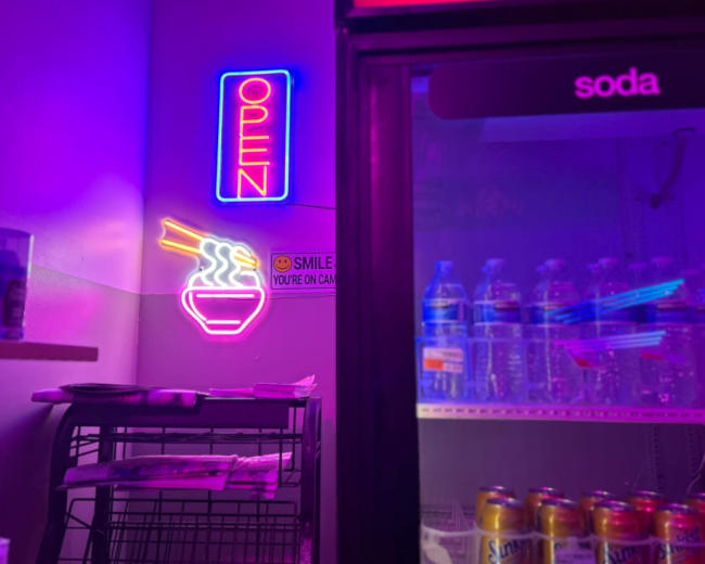 A brightly lit neon sign that says "OPEN" hangs next to a bowl of noodles, while shelves stocked with bottled water and soda cans are visible in a nearby refrigerator.