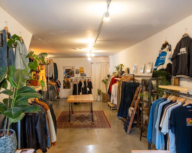 The image shows a clothing store interior featuring racks of various apparel, a wooden table in the center, and potted plants throughout.