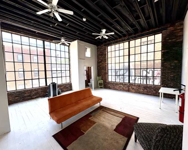 The image shows a spacious room with large windows, exposed brick walls, and minimal furnishings, including a vintage orange sofa and a small white table.