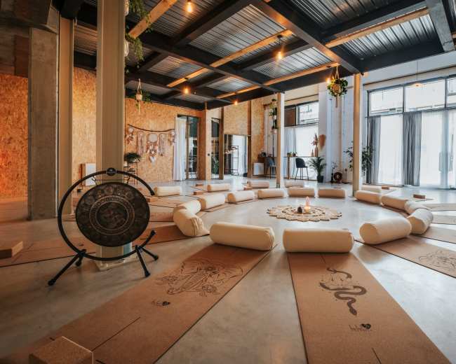 The spacious room features yoga mats arranged in a circular formation around a decorative centerpiece, with natural light filtering through large windows.