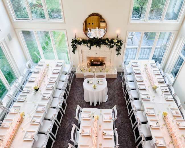 The image features a spacious dining area set with two long tables arranged in a U-shape, decorated with flowers and candles, and a fireplace adorned with greenery.
