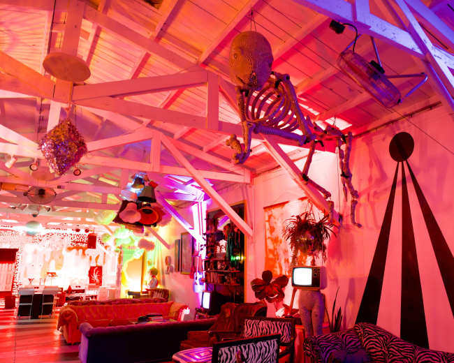 The interior of a colorful, artistic lounge features a skeleton suspended from the ceiling, surrounded by various eclectic decorations and vibrant lighting.