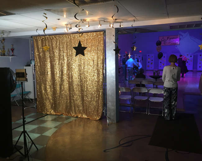 A glittering gold curtain stands at the entrance to a dimly lit event space decorated for a celebration, with people mingling in the background.